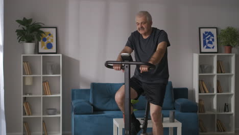 home-fitness-and-healthy-lifestyle-of-middle-aged-man-is-training-on-stationary-bike-in-apartment-cardio-workout-for-keeping-good-physical-condition
